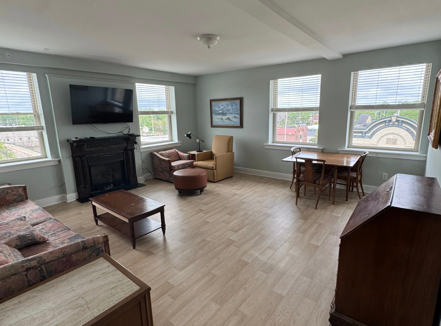 A bright and spacious independent-living penthouse apartment at Black Hawk Senior Residence, showcasing a large living room and dining area with light wood-toned flooring and soft seafoam green walls. The space features three large windows with white blinds overlooking downtown, letting in abundant natural daylight. The living area includes a floral print sofa, a dark fireplace mantle with a flat-screen TV mounted above, a wooden coffee table, and comfortable armchairs with a leather ottoman. To the right, a wooden dining table with chairs sits near an elegant vintage writing desk.
