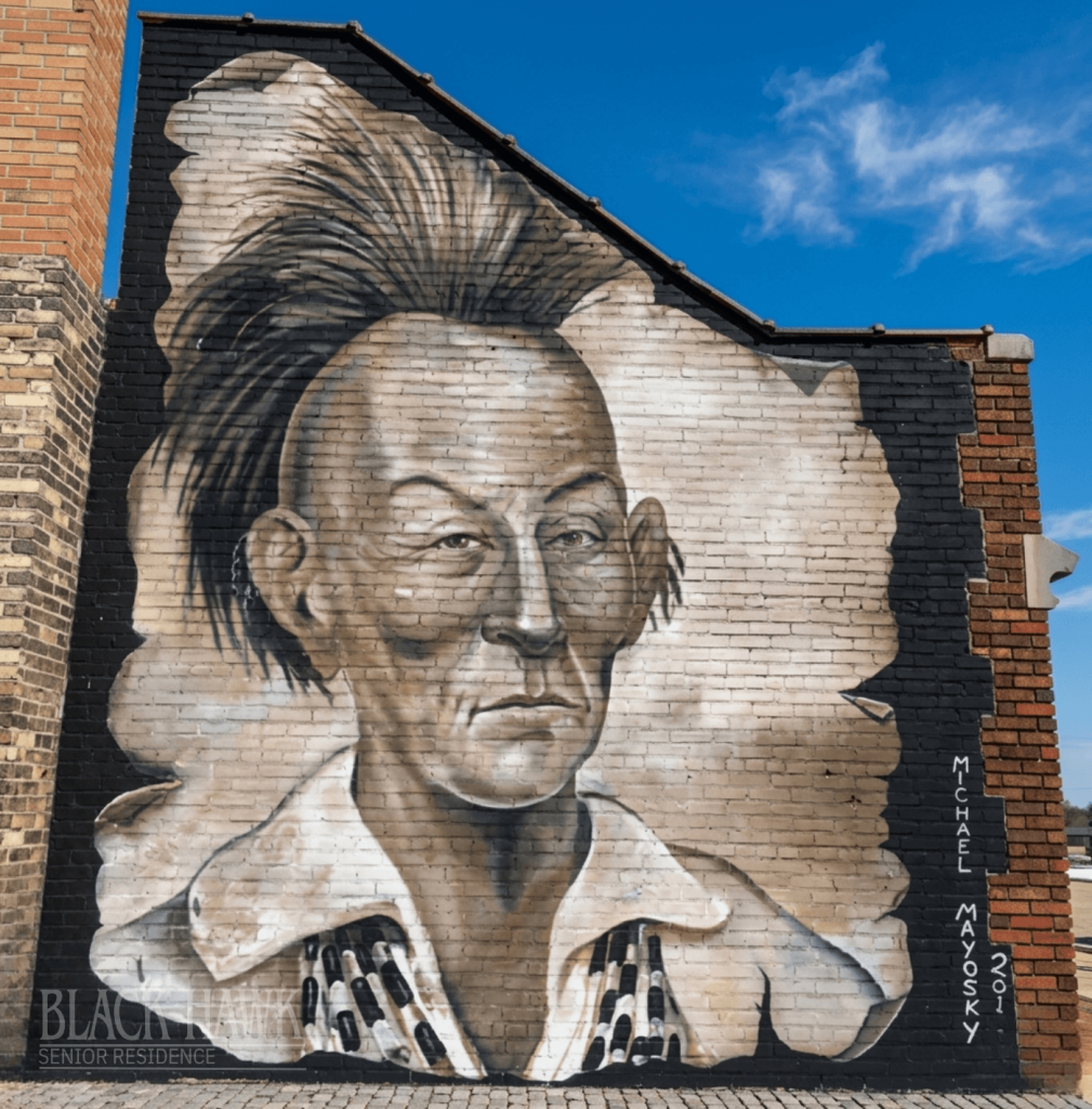 A large, realistic mural of Native American leader Black Hawk, with a Mohawk hairstyle and a beaded choker, painted on the exterior brick wall of a building.