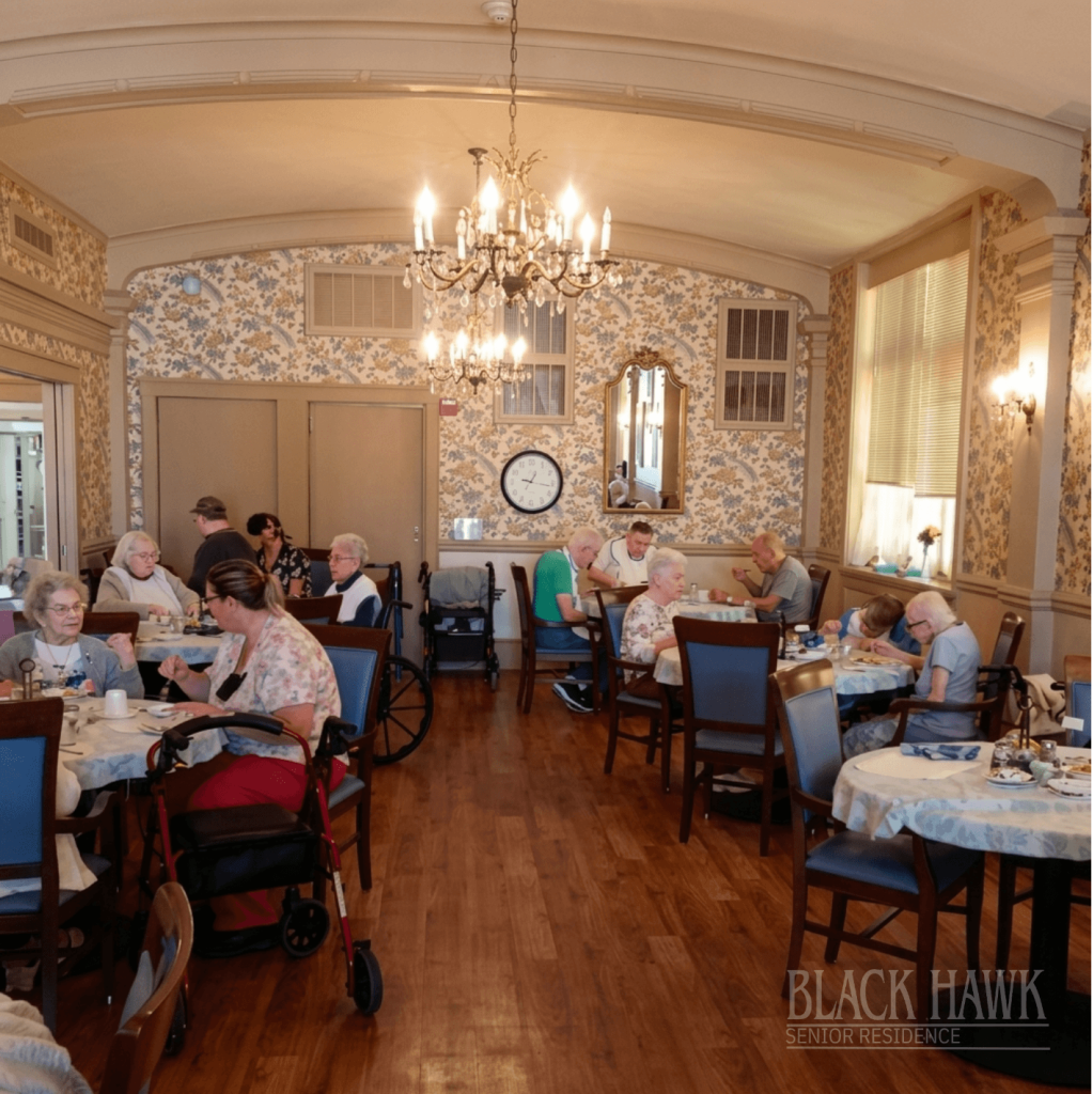 A wide interior view of an elegant dining hall with a coffered ceiling and crystal chandelier, where senior residents are seated at tables for dinner.