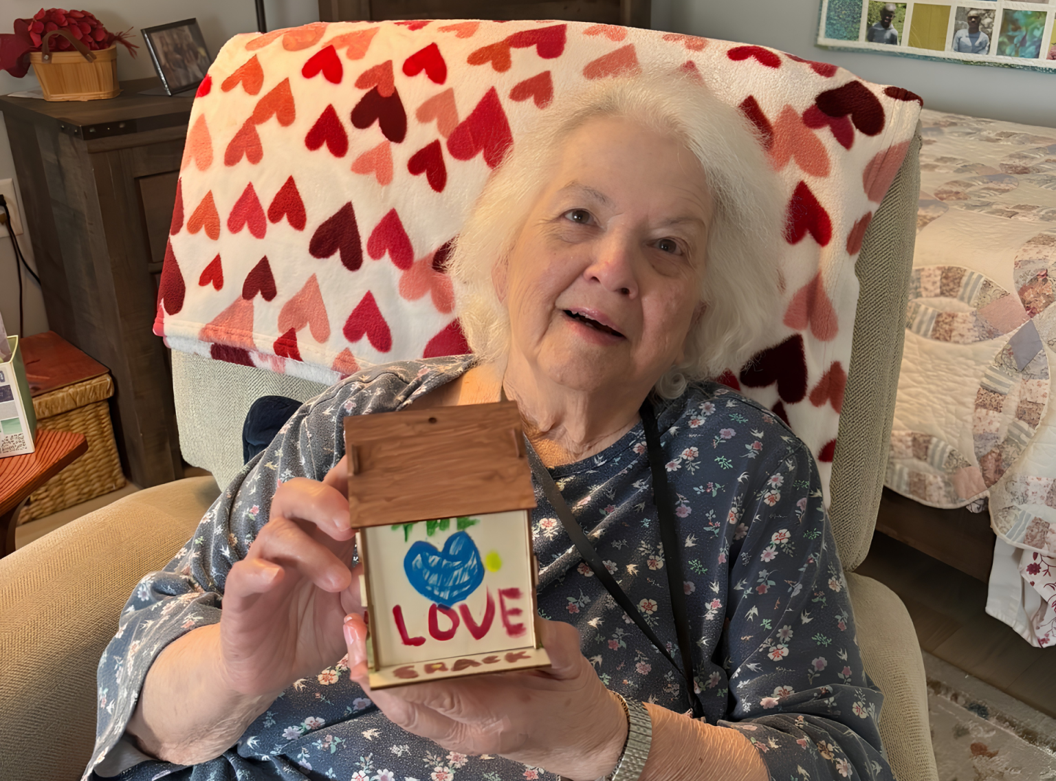 A heartwarming close-up of an elderly female resident with white hair smiling warmly at Black Hawk Senior Residence. She is sitting in a comfortable armchair with a red and pink heart-patterned blanket draped behind her. In her hands, she proudly holds up a small, handmade wooden craft house decorated with a blue painted heart and the word 'LOVE' written in red letters. She wears a floral print blue top and a silver watch on her wrist, creating a cozy and joyful atmosphere.