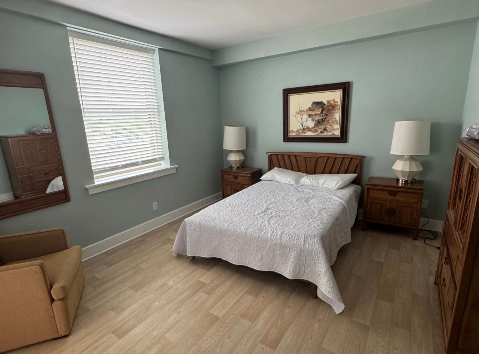 A bright and spacious resident bedroom at Black Hawk Senior Residence, featuring a beautifully made double bed with white textured linens and a warm wooden headboard. Matching wooden nightstands with white geometric lamps sit on either side of the bed under a framed landscape painting. The room features smooth, light wood-toned flooring, light seafoam green walls, a comfortable upholstered armchair in the corner, and a large window with white blinds that lets in soft, natural daylight.