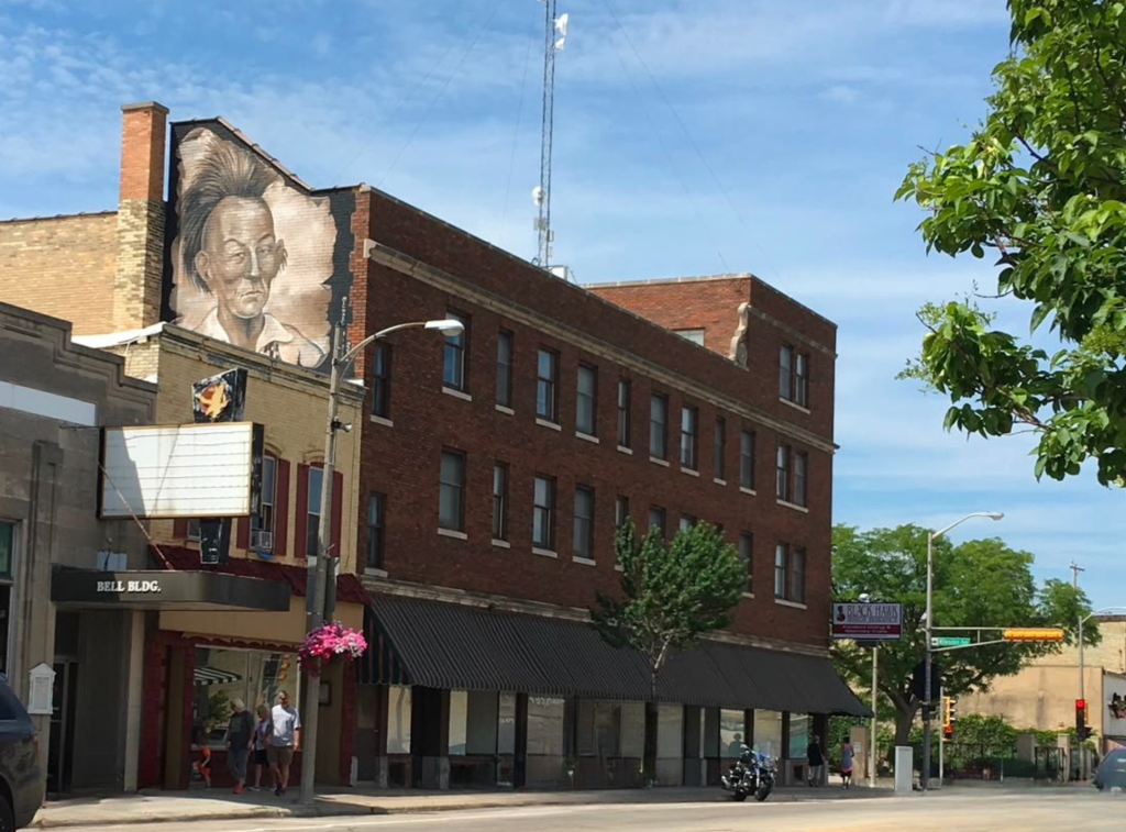 Exterior view of the historic brick building of Black Hawk Senior Residence, featuring a prominent Native American portrait mural on the upper facade, street-level awnings, and a sunny sky.